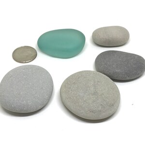 May include: A collection of smooth, rounded stones in various shades of gray and a single piece of sea glass in a soft teal color. A coin is included for size comparison. The stones vary in size and shape, with a matte finish.