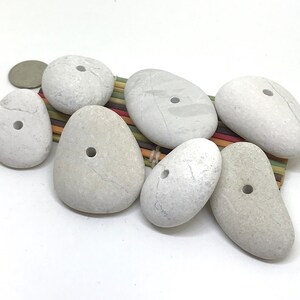 May include: Seven white smooth stones with holes drilled through the center. The stones are arranged on a woven mat with a light brown background.