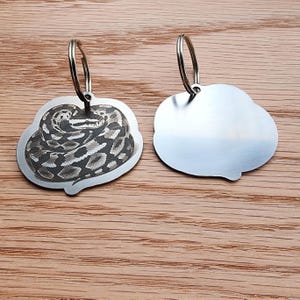 Op de afbeelding: Twee metalen hondenpenningen met een slangenontwerp. E&eacute;n penning heeft een gedetailleerde gravure van een opgerolde slang, terwijl de andere leeg is. Beide penningen hebben een zilverkleurige afwerking en een ronde ring voor bevestiging. De penningen worden op een houten oppervlak getoond.