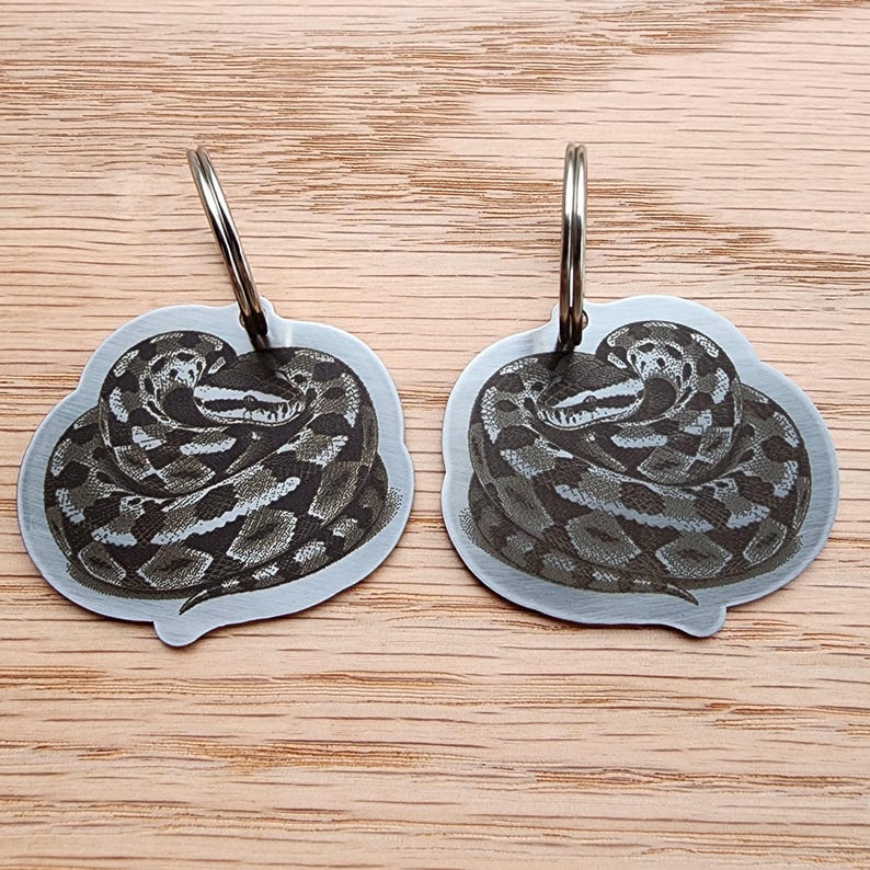 Op de afbeelding: Twee metalen sleutelhangers met een gedetailleerde illustratie van een opgerolde slang. De slang is afgebeeld in grijstinten en bruin, met een textuurpatroon. Elke sleutelhanger heeft een zilverkleurige ring aan de bovenkant bevestigd. De achtergrond is een lichtbruin houten oppervlak.