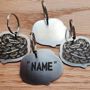 May include: Four silver-colored keychains with a snake design. Three keychains feature a coiled snake illustration. One keychain has the word "NAME" in black text. All keychains have a silver key ring attached.