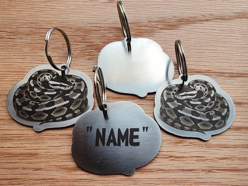 Op de afbeelding: Vier zilverkleurige sleutelhangers met een slangenontwerp. Drie sleutelhangers hebben een illustratie van een opgerolde slang. E&eacute;n sleutelhanger heeft het woord "NAME" in zwarte tekst. Alle sleutelhangers hebben een zilveren sleutelring.