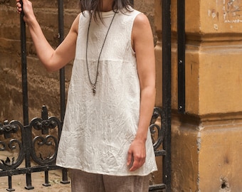 May include: A woman wearing a white sleeveless top with a long pendant necklace and brown linen pants.