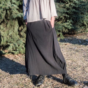 Op de afbeelding: Een vrouw draagt een lichtbeige, loszittende blouse met lange mouwen en een geplooide taille en een zwarte, zwierige maxi-rok. Ze staat voor een groene naaldbomen.