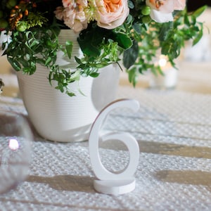 Può includere: Un numero di tavolo bianco, il sei, è posto su una tovaglia bianca, con una composizione floreale sullo sfondo. I fiori sono in un vaso bianco. La tavola è apparecchiata con bicchieri e un cestino.