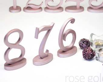 May include: Rose gold wedding table numbers 1 through 8. The numbers are made of wood and have a cursive font. They are displayed on a white background.