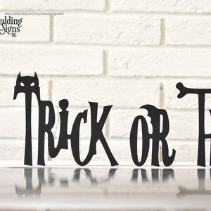 May include: Black silhouette cutout of the phrase "Trick or Treat" with a small bat silhouette above the word "Trick". The cutout is displayed on a white brick wall background.
