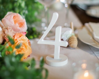 May include: A white wooden table number '4' sits on a white tablecloth with a bouquet of peach and cream colored flowers in the foreground.