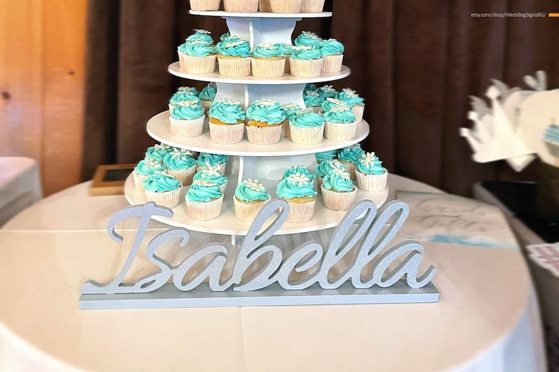 Custom wooden name sign in white finish beside tiered blue cake, elegant Quinceañera mis quince centerpiece. A four-tier cupcake stand with teal cupcakes decorated with white snowflakes. The stand is decorated with a silver glitter sign that reads "Isabella".