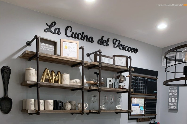 Puede incluir: Un conjunto de estantes de metal negro con estantes de madera, decorados con varios art&iacute;culos, incluyendo un letrero que dice "La Cucina del Vescovo", un letrero "AM" dorado y un letrero "PM" negro.