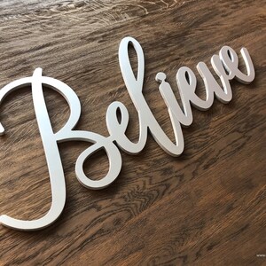 Könnte beinhalten: Ein silbernes Metallschild mit dem Wort "Believe" in kursivschrift. Das Schild befindet sich auf einem hölzernen Hintergrund.