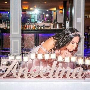 Custom wooden name sign in gold finish at candlelit Quinceañera reception table centerpiece. A young woman in a pink dress blows out candles on a table with the name "Angelina" spelled out in large, glittery letters. The candles are in glass holders and are surrounded by smoke.