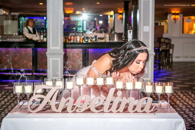 Custom wooden name sign in gold finish at candlelit Quinceañera reception table centerpiece. A young woman in a pink dress blows out candles on a table with the name "Angelina" spelled out in large, glittery letters. The candles are in glass holders and are surrounded by smoke.