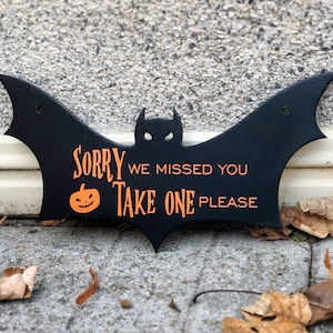 May include: Black bat-shaped sign with the text "Sorry we missed you Take one please" in orange lettering. A small orange pumpkin with a jack-o'-lantern face is next to the text "Sorry".