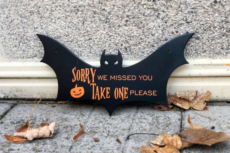 May include: Black bat-shaped sign with the text "Sorry we missed you Take one please" in orange lettering. A small orange pumpkin with a jack-o'-lantern face is next to the text "Sorry".