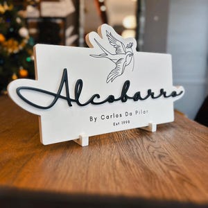 May include: A white wooden sign with the name "Alcobarra" in black cursive lettering. A black line drawing of a bird holding a sprig is above the name. The text "By Carlos Da Pilar Est 1998" is below the name. The sign is on a wooden surface.