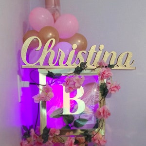 Personalized name sign in purple neon light setting, princess theme Quinceañera birthday centerpiece. A gold glitter name sign with the name "Christina" on it. The sign is decorated with pink flowers and greenery. A light-up box with the letter "B" inside is below the sign.