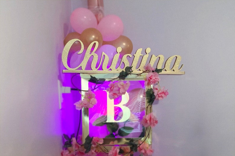 Personalized name sign in purple neon light setting, princess theme Quinceañera birthday centerpiece. A gold glitter name sign with the name "Christina" on it. The sign is decorated with pink flowers and greenery. A light-up box with the letter "B" inside is below the sign.