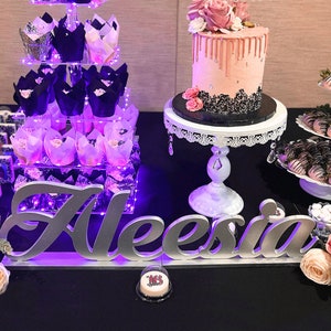 Custom wooden name sign in pink and purple glitter finish, glamorous Quinceañera gift centerpiece.  A dessert table with a pink and black drip cake, a tiered stand with cupcakes, and a sign that says "Aleesia". The table is decorated with flowers and chocolate-covered strawberries.