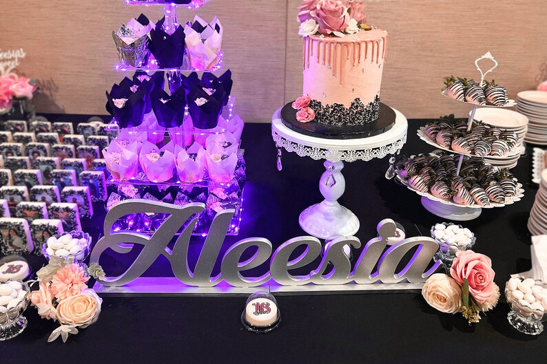 Custom wooden name sign in pink and purple glitter finish, glamorous Quinceañera gift centerpiece.  A dessert table with a pink and black drip cake, a tiered stand with cupcakes, and a sign that says "Aleesia". The table is decorated with flowers and chocolate-covered strawberries.