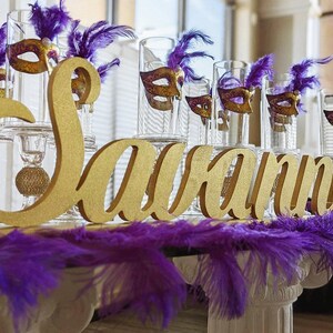 Large personalized name sign in gold finish at Quinceañera sweetheart table, mis quince gift keepsake. Gold glitter letters spelling "Savannah" with purple feathers and glass candle holders with gold masquerade masks.