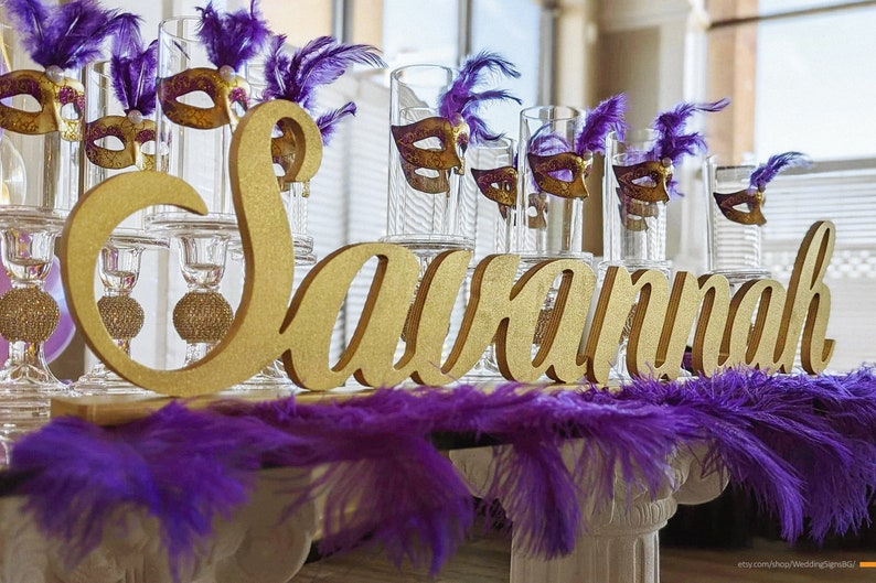 Large personalized name sign in gold finish at Quinceañera sweetheart table, mis quince gift keepsake. Gold glitter letters spelling "Savannah" with purple feathers and glass candle holders with gold masquerade masks.
