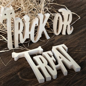May include: Wooden cut-out letters spelling "Trick or Treat" in a whimsical font. The letters are light brown and have a slightly distressed look. The letters are arranged on a dark brown wooden surface.