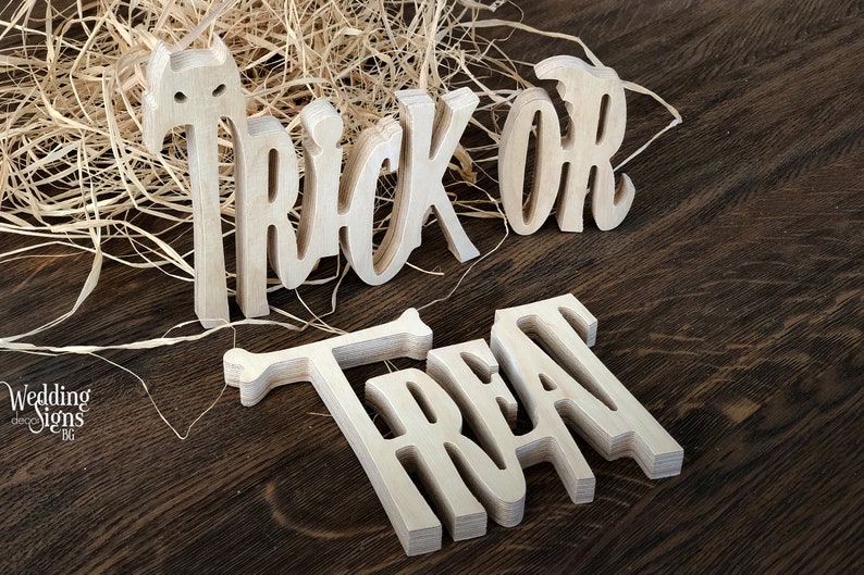 May include: Wooden cut-out letters spelling "Trick or Treat" in a whimsical font. The letters are light brown and have a slightly distressed look. The letters are arranged on a dark brown wooden surface.