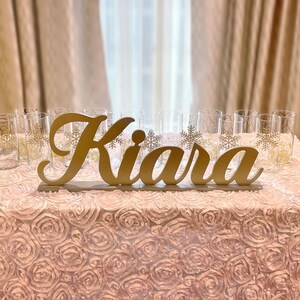 Personalized name sign in gold finish on sequin tablecloth, elegant Quinceañera mis quince gift decor. A gold wooden sign with the name "Kiara" on a pink, ruffled table cloth. There are clear glass cups with gold snowflake designs on the table.