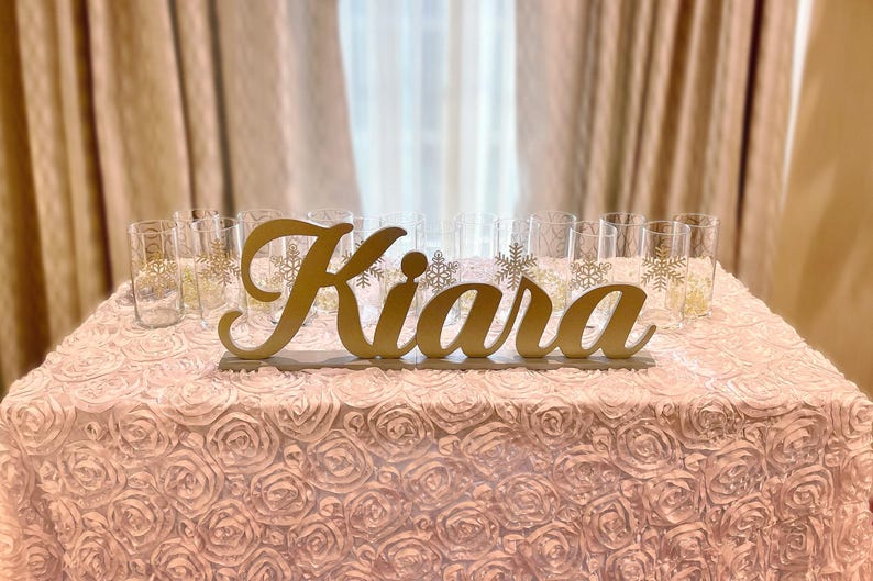 Personalized name sign in gold finish on sequin tablecloth, elegant Quinceañera mis quince gift decor. A gold wooden sign with the name "Kiara" on a pink, ruffled table cloth. There are clear glass cups with gold snowflake designs on the table.