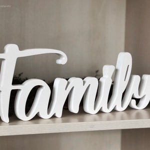 Może przedstawiać: Biały drewniany znak z napisem "Family" w stylu ozdobnym. Znak stoi na drewnianej p&oacute;łce.