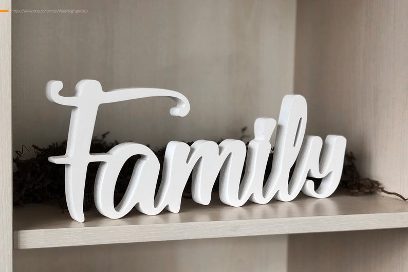 Może przedstawiać: Biały drewniany znak z napisem "Family" w stylu ozdobnym. Znak stoi na drewnianej p&oacute;łce.