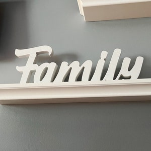 Może przedstawiać: Biała drewniana p&oacute;łka z napisem "Family" w białych literach. P&oacute;łka jest zamontowana na szarej ścianie.