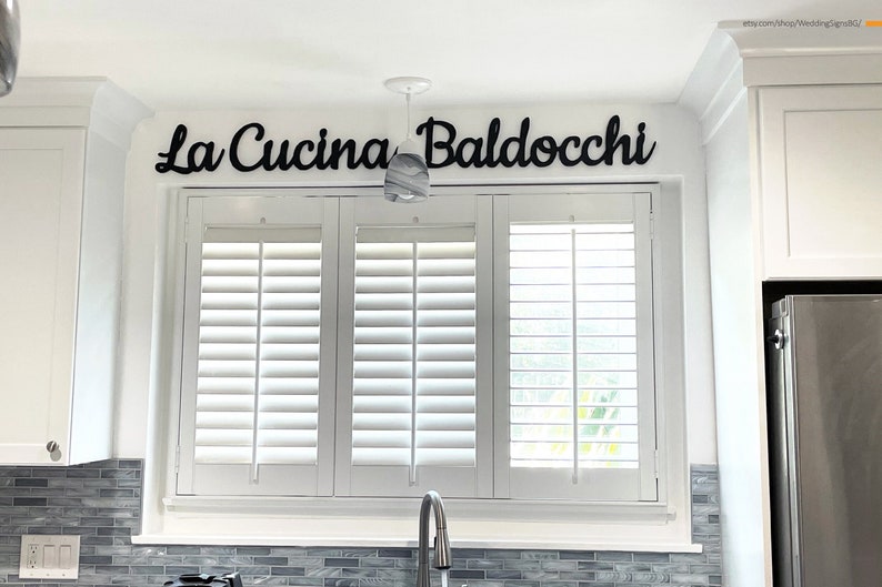 Puede incluir: Una cocina blanca con una ventana que tiene persianas blancas de madera. Sobre la ventana hay un letrero de madera negro que dice "La Cucina Baldocchi".