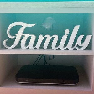 Może przedstawiać: Biała drewniana szafka z białym napisem "Family" na g&oacute;rnej p&oacute;łce. Na dolnej p&oacute;łce znajduje się czarny elektroniczny sprzęt z napisem "Sky".