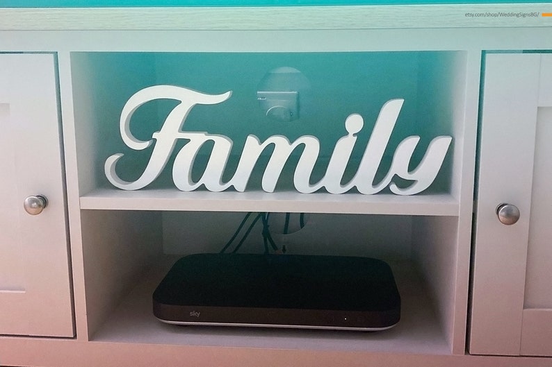 Może przedstawiać: Biała drewniana szafka z białym napisem "Family" na g&oacute;rnej p&oacute;łce. Na dolnej p&oacute;łce znajduje się czarny elektroniczny sprzęt z napisem "Sky".
