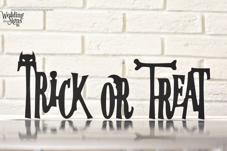 May include: Black silhouette cutout of the phrase "Trick or Treat" with a small bat silhouette above the word "Trick". The cutout is displayed on a white brick wall background.