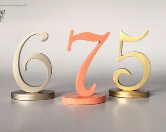 May include: Three wooden table numbers, 6, 7, and 5, in gold, coral, and silver. The numbers are in a cursive font and are on small stands.