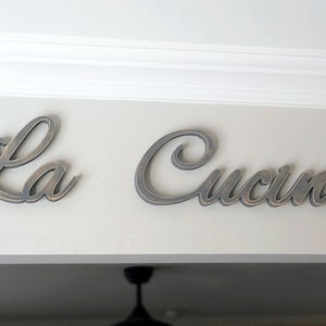 Puede incluir: Primer plano de un letrero de madera que dice "La Cucina" en una fuente cursiva. El letrero est&aacute; pintado en un color gris desgastado y est&aacute; montado en una pared blanca.