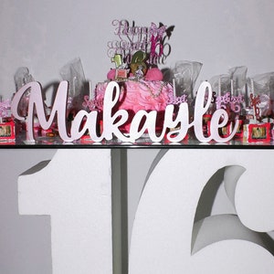 Multiple custom wooden name signs in silver finish displayed together, Quinceañera gift centerpiece options. A pink and white cake with a pink and white cake topper that says "Happy Sweet 16" and a pink and white sign that says "Makayle". The cake is surrounded by small pink and white cupcakes and other decorations.