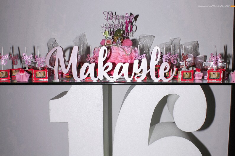 Multiple custom wooden name signs in silver finish displayed together, Quinceañera gift centerpiece options. A pink and white cake with a pink and white cake topper that says "Happy Sweet 16" and a pink and white sign that says "Makayle". The cake is surrounded by small pink and white cupcakes and other decorations.