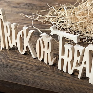 May include: Wooden sign with the words "Trick or Treat" in a decorative font. The sign is made of light brown wood and is sitting on a dark brown wooden surface.