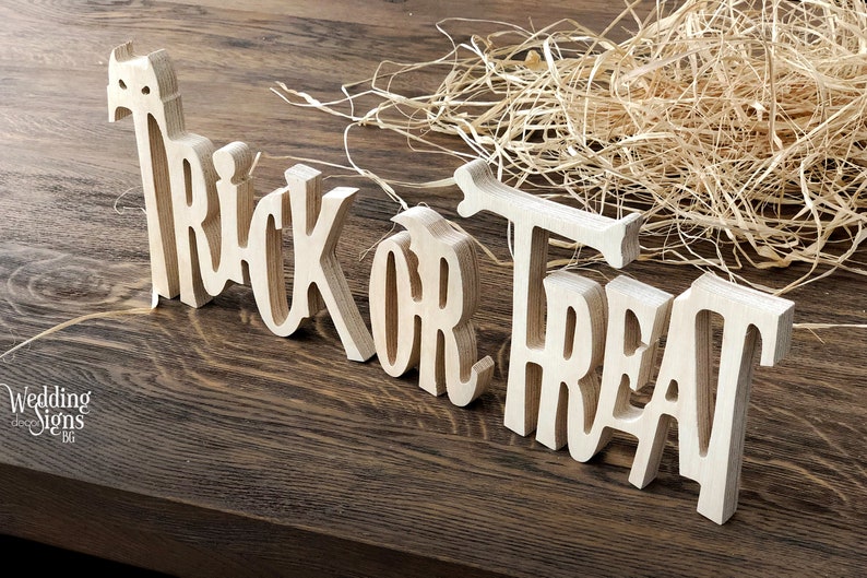 May include: Wooden sign with the words "Trick or Treat" in a decorative font. The sign is made of light brown wood and is sitting on a dark brown wooden surface.