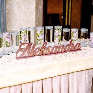 Multiple personalized name signs in gold finish on long decorated table, Quinceañera banquet centerpieces. A table decorated with clear glass cylinders filled with water and purple flowers. The table is covered with a white tablecloth and has a wooden sign that reads "Aleksandra".