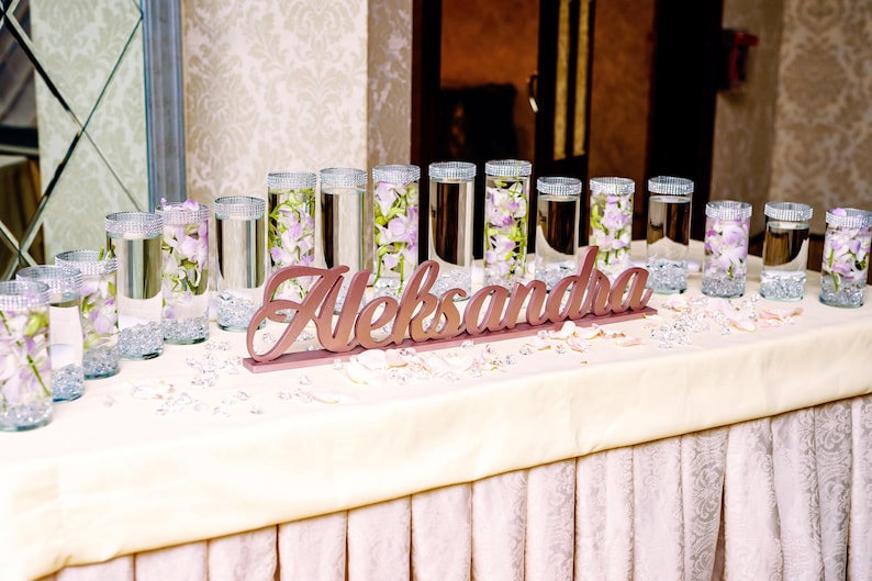 Multiple personalized name signs in gold finish on long decorated table, Quinceañera banquet centerpieces. A table decorated with clear glass cylinders filled with water and purple flowers. The table is covered with a white tablecloth and has a wooden sign that reads "Aleksandra".
