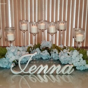Custom wooden name sign in silver finish on elegant white tablecloth, mis quince party centerpiece. A silver glitter name sign spelling "Jenna" is surrounded by a garland of light blue flowers and green leaves.  The sign is placed in front of a row of clear glass candle holders with white candles.