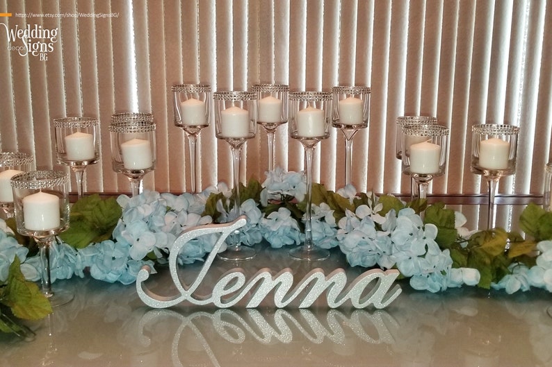 Custom wooden name sign in silver finish on elegant white tablecloth, mis quince party centerpiece. A silver glitter name sign spelling "Jenna" is surrounded by a garland of light blue flowers and green leaves.  The sign is placed in front of a row of clear glass candle holders with white candles.