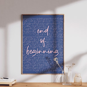 May include: A framed print with a navy blue background and a handwritten script. The words "end of beginning" are in pink cursive. The print is displayed on a wooden surface with decorative items.