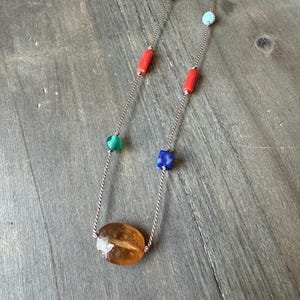 May include: A long necklace with a silver chain and various colorful beads. The necklace features an oval, amber-colored stone pendant and is accented with red, green, blue, and turquoise beads. The necklace is displayed on a wooden surface.
