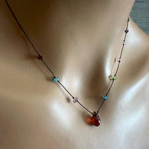 Puede incluir: Delicado collar con un cordón negro adornado con cuentas de colores. El collar tiene un colgante en forma de lágrima de color ámbar. Las cuentas son en tonos de rojo, azul, morado, verde y transparente.